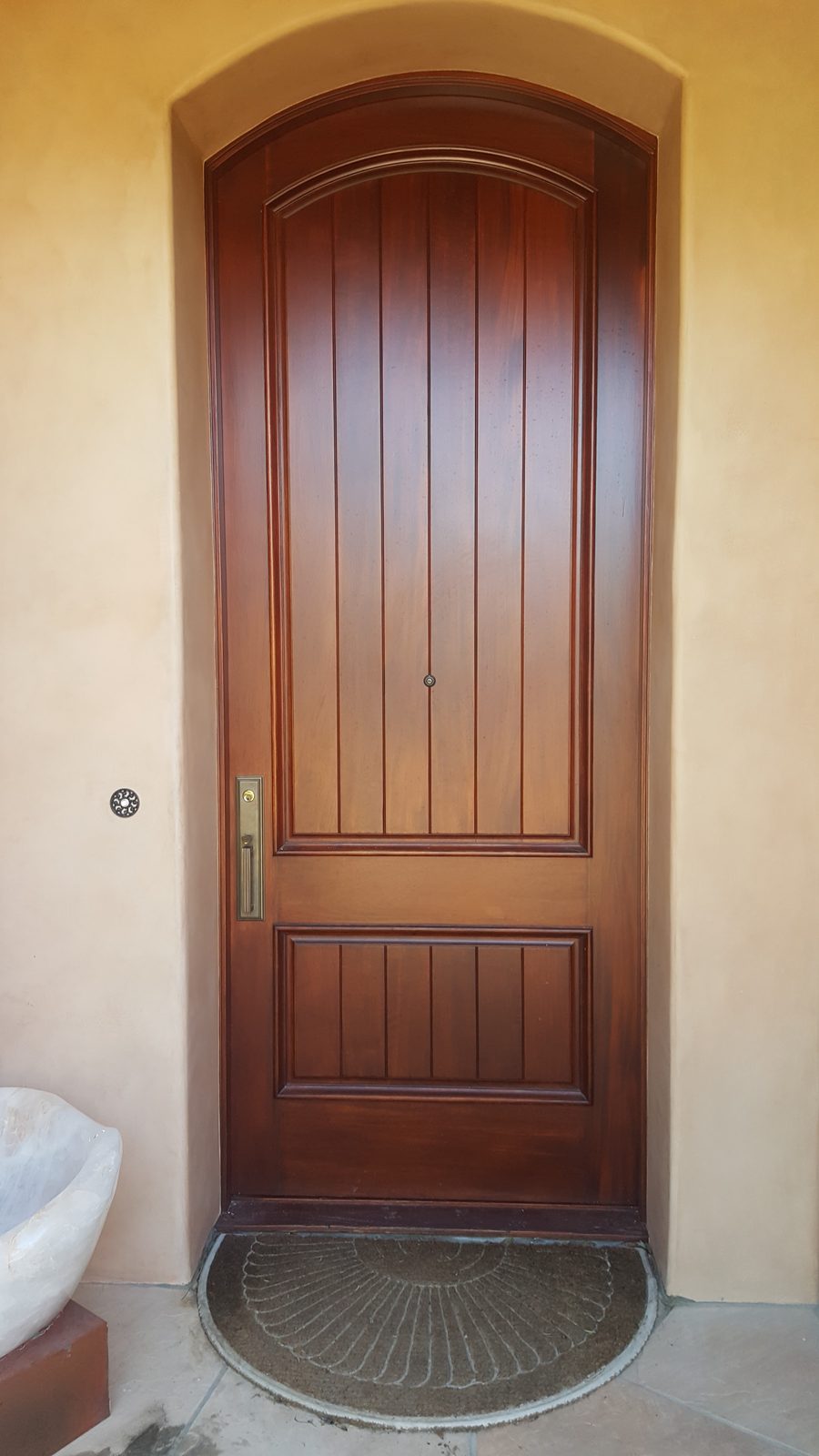 An arched, plank-paneled front door in a deep reddish stained finish, set in a curved stucco entry.