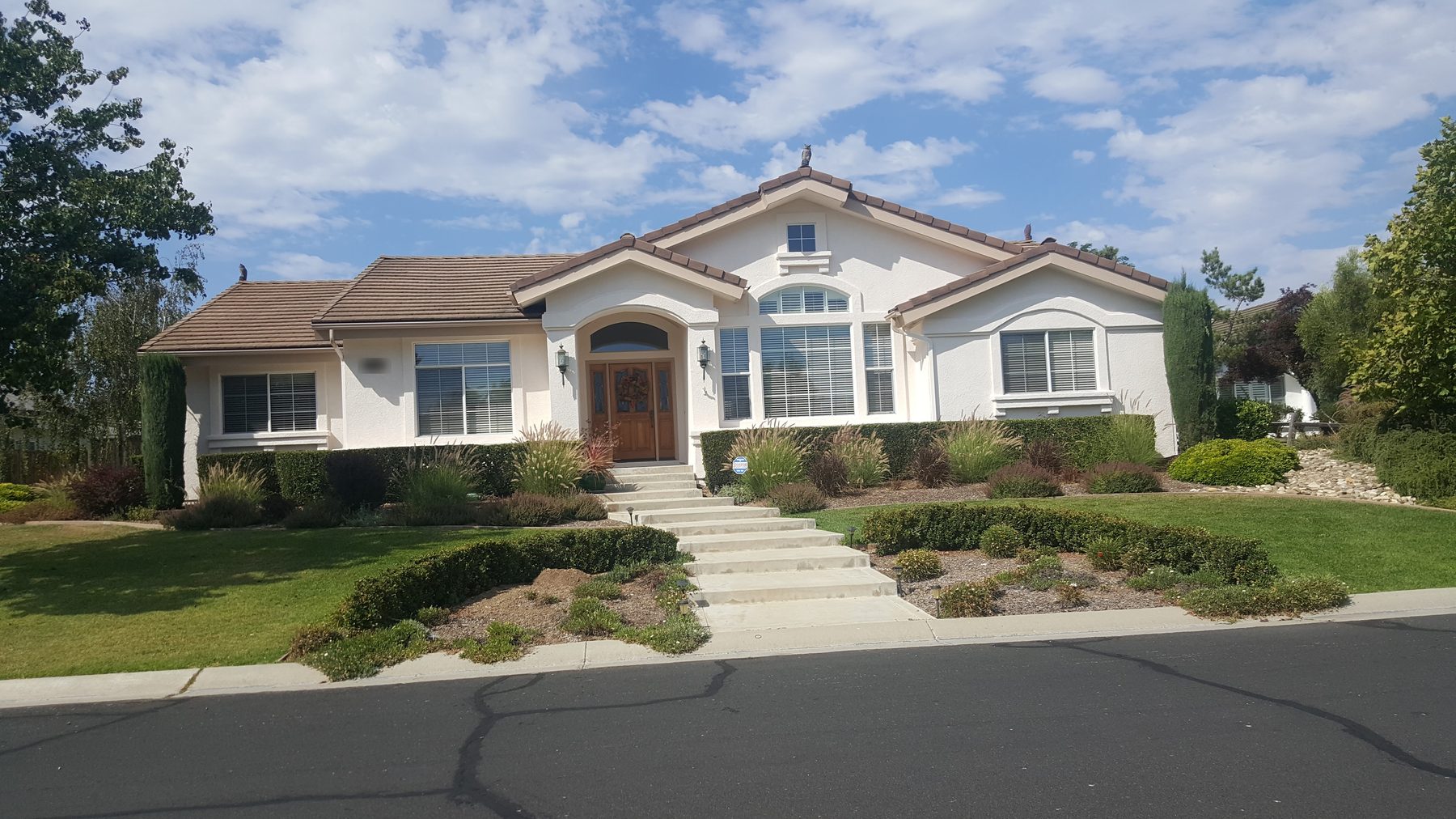 A single-story cream stucco house with a tile roof, arched entry and wide front steps, behind a green lawn and trimmed hedges.