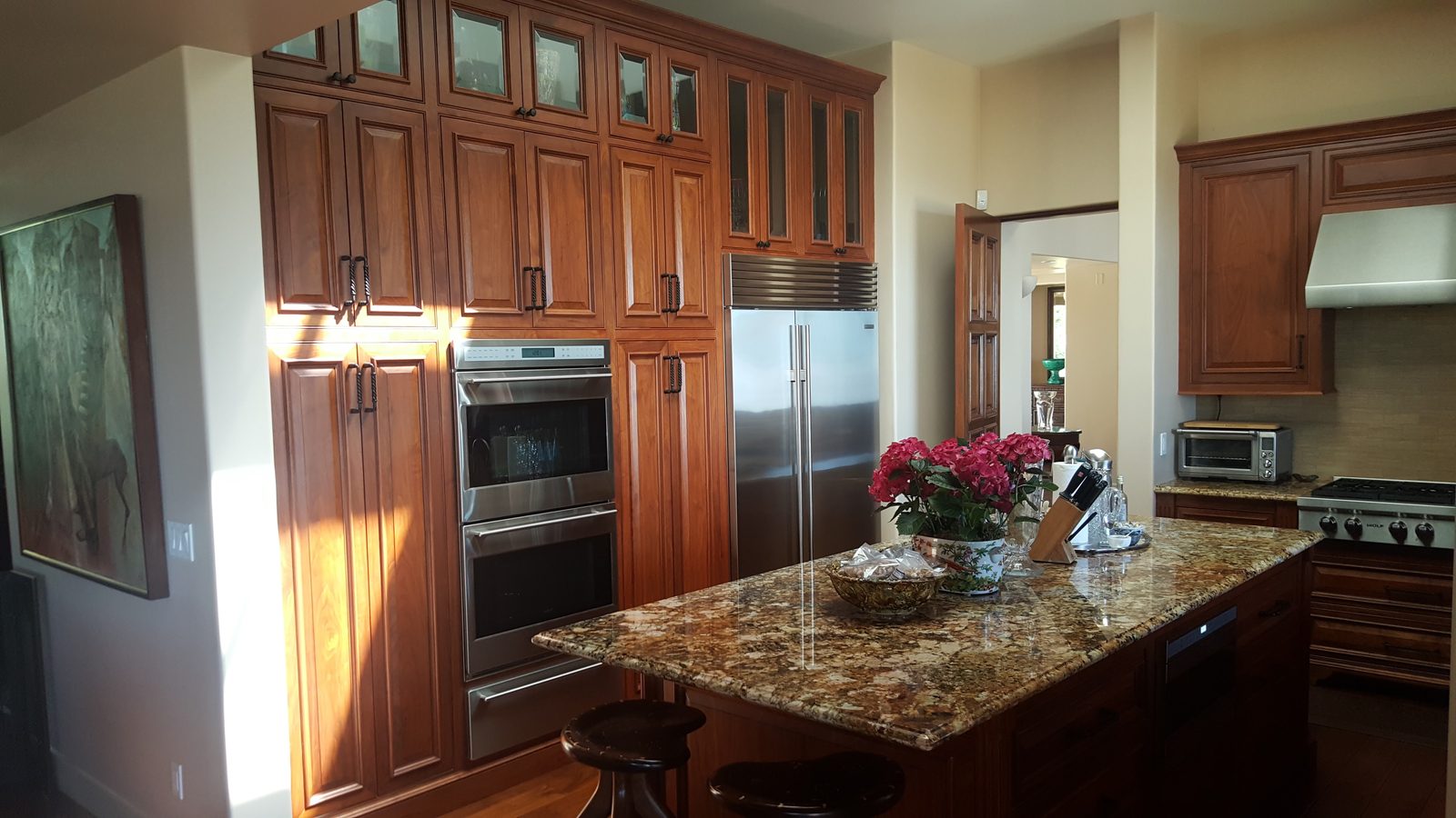A tall wall of reddish-brown stained kitchen cabinets with glass-front uppers around stainless double ovens and a refrigerator, beside a granite island.