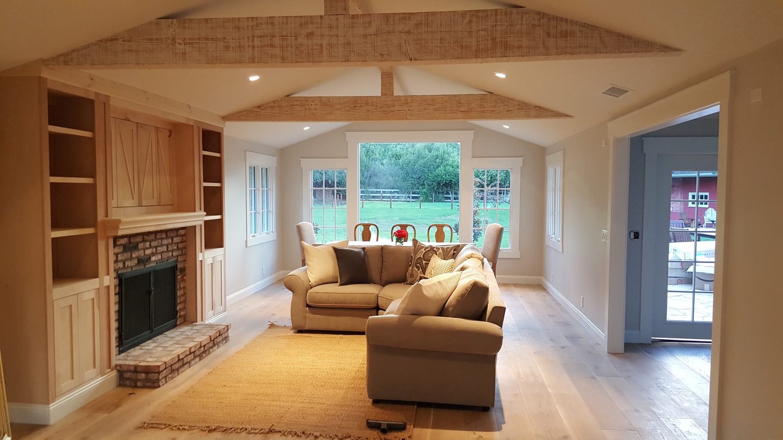 A living room with new built-in shelves and cabinets in bare, unfinished wood around a brick fireplace, under exposed rough-sawn beams.